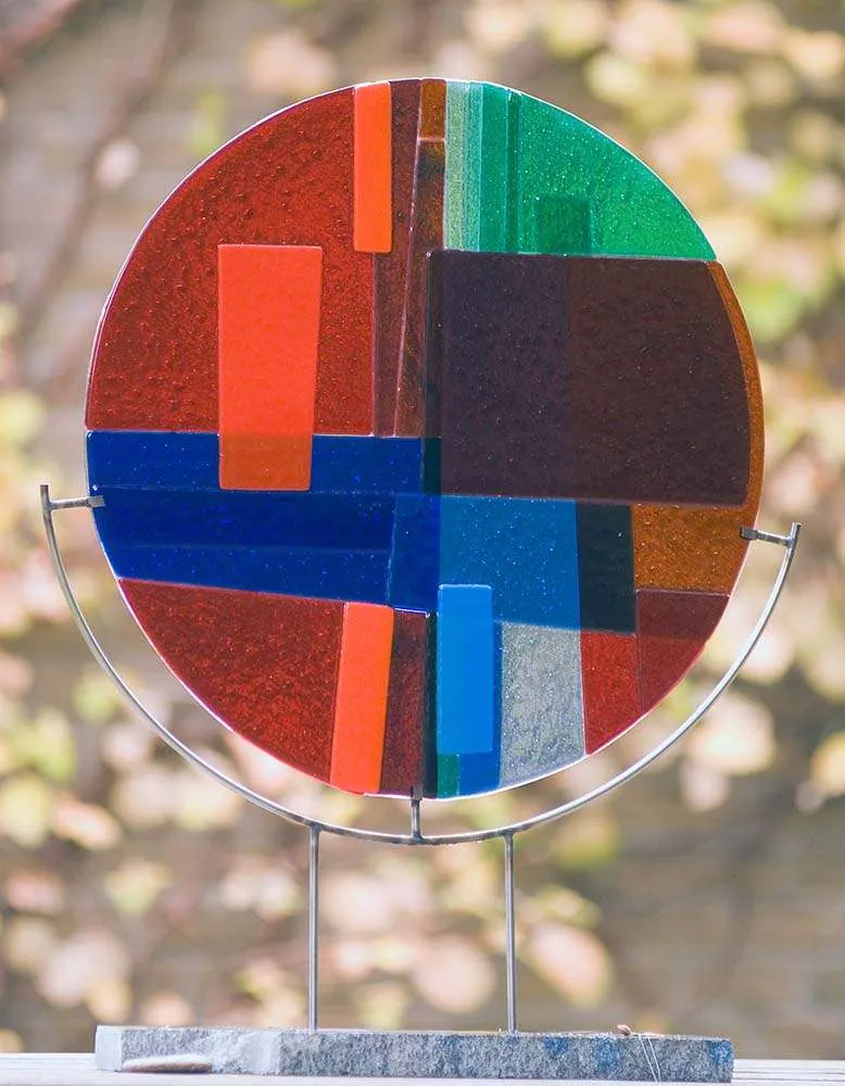 Rond glaskunstwerk in rood, blauw en groen, op een standaard in de tuin